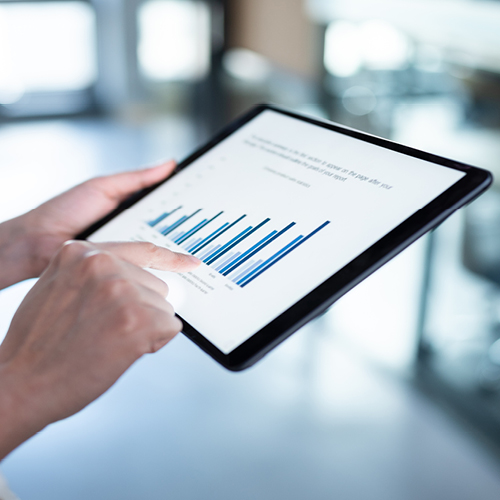 Hand holding a tablet displaying a bar chart, pointing at data on the screen in a bright, blurred office setting.