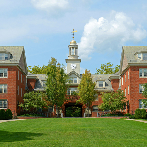 Red brick academic building with a central clock tower, arched passageway, and green lawn in front under a blue sky.