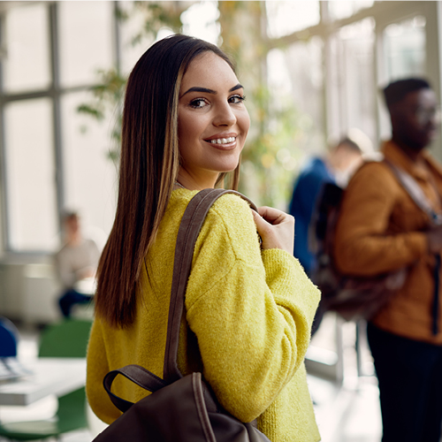 Smiling person in a yellow sweater with a backpack turning toward the camera in a bright campus hallway.