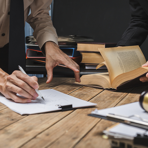 Hands writing on a clipboard at a wooden table while someone flips through a book, with stacked books in the background.