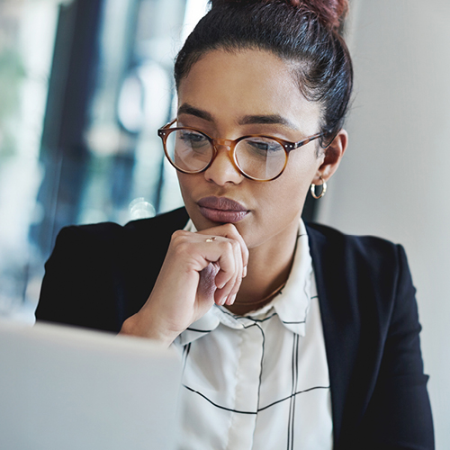 Person in casual business attire and glasses looking at a laptop with hand on chin in a modern glass office.