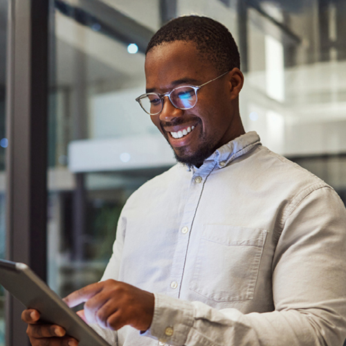 Person wearing glasses, smiling and pointing at a tablet in a modern glass office with interior lighting.
