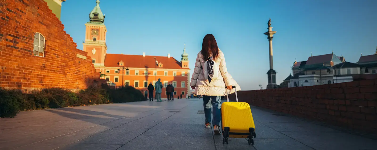 A woman with a white coat pulls a yellow suitcase on wheels next to a brick wall. 