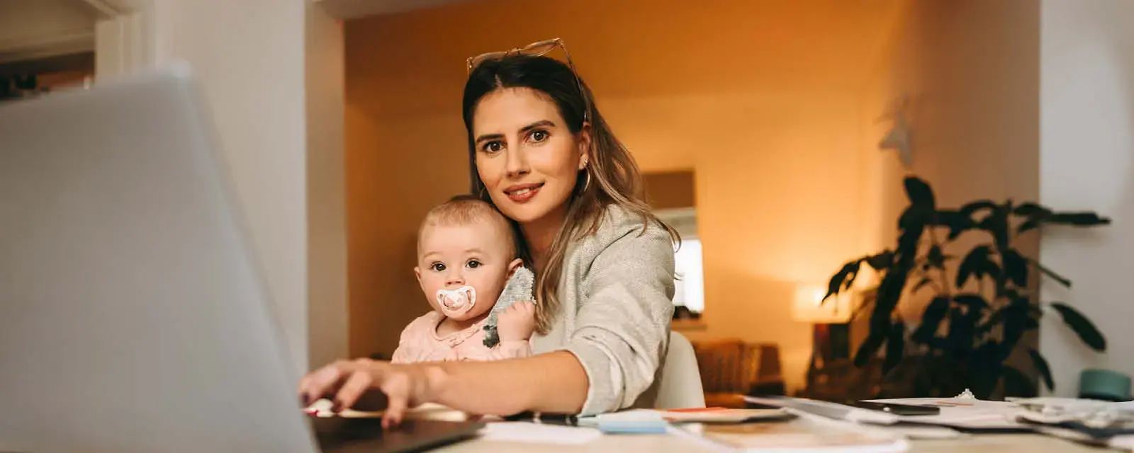 Single mom typing on a laptop while working in her home office. Multitasking mom working on a new creative project at her desk. Female interior designer carrying her baby on her laps.