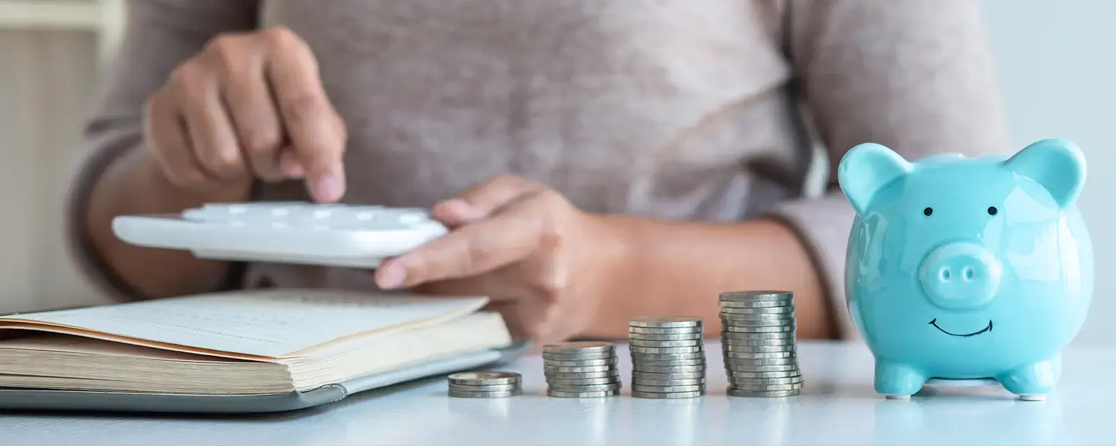Close-up of a person using a calculator with a piggy bank and coins in the foreground. 