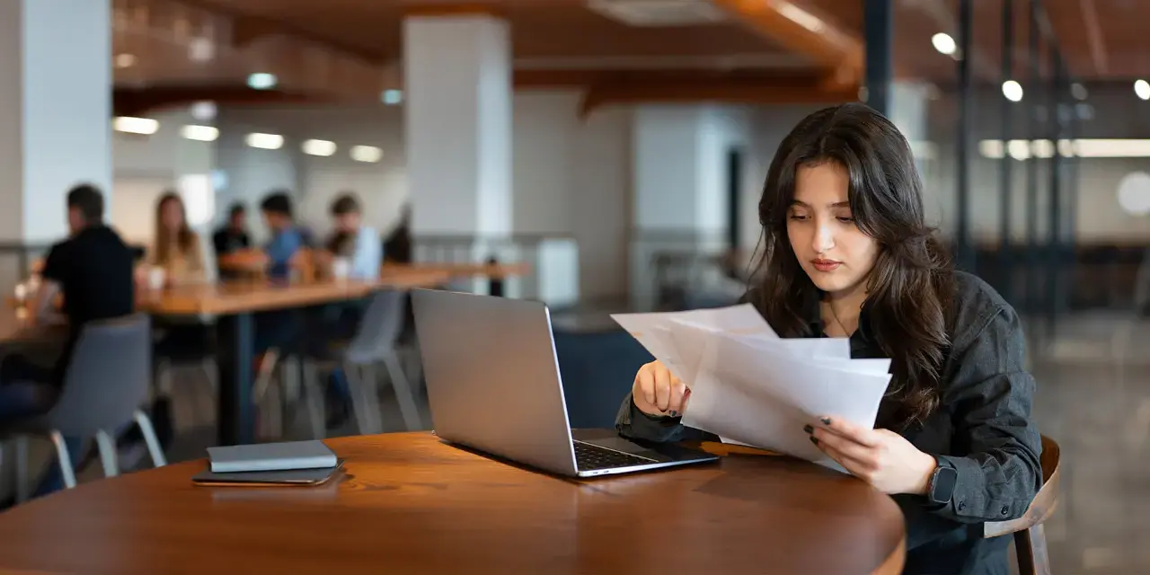 Young woman works with laptop in cafe and examines financial report papers.