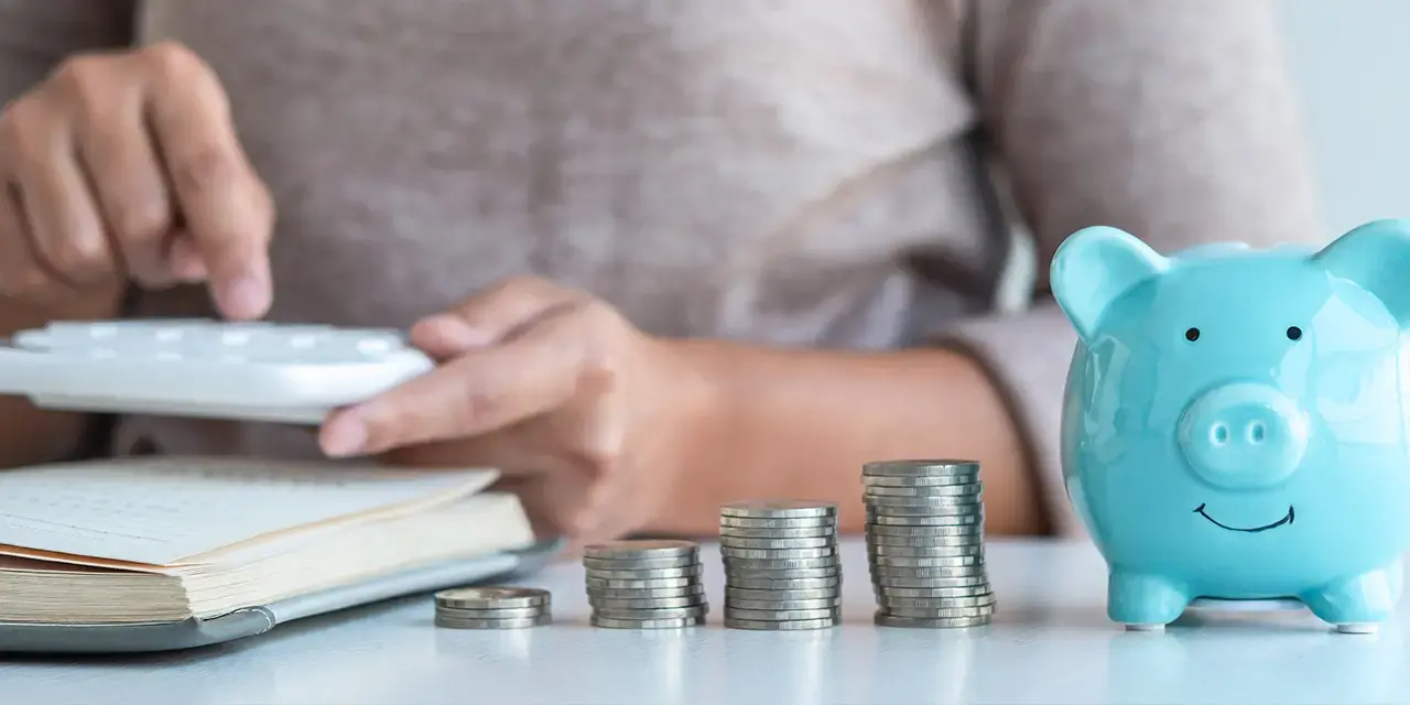 Close-up of a person using a calculator with a piggy bank and coins in the foreground. 