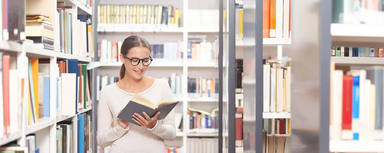 A young woman with glasses reading a book in a modern library. Bookshelves filled with colorful books in the background. Concept of studying, education, learning, knowledge, and academic lifestyle