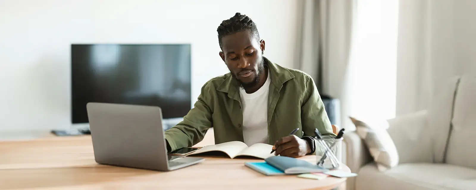 "Focused African-American man sitting at desk working on laptop and taking notes in notebook at home.   "