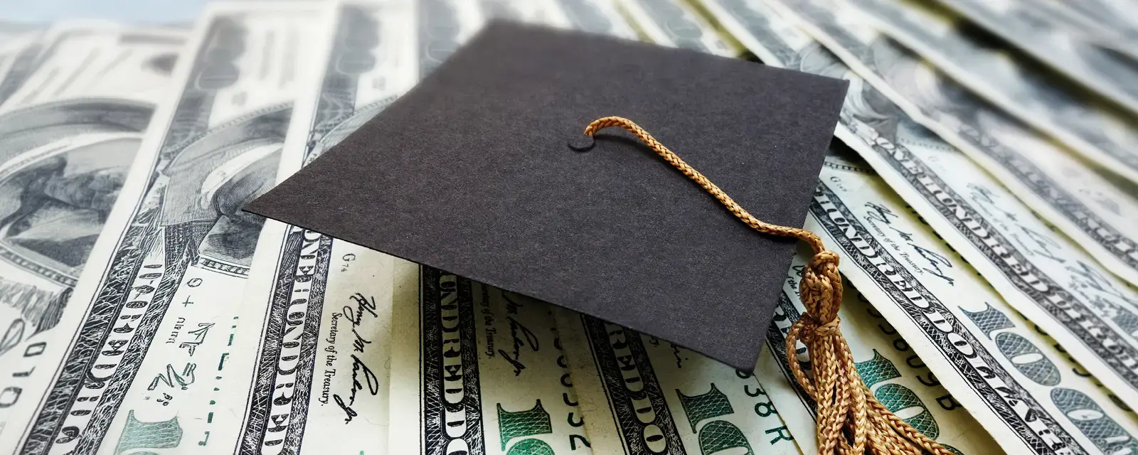 Close up of a black graduation cap, or mortaboard, with gold tassel on top of hundred dollar bills
