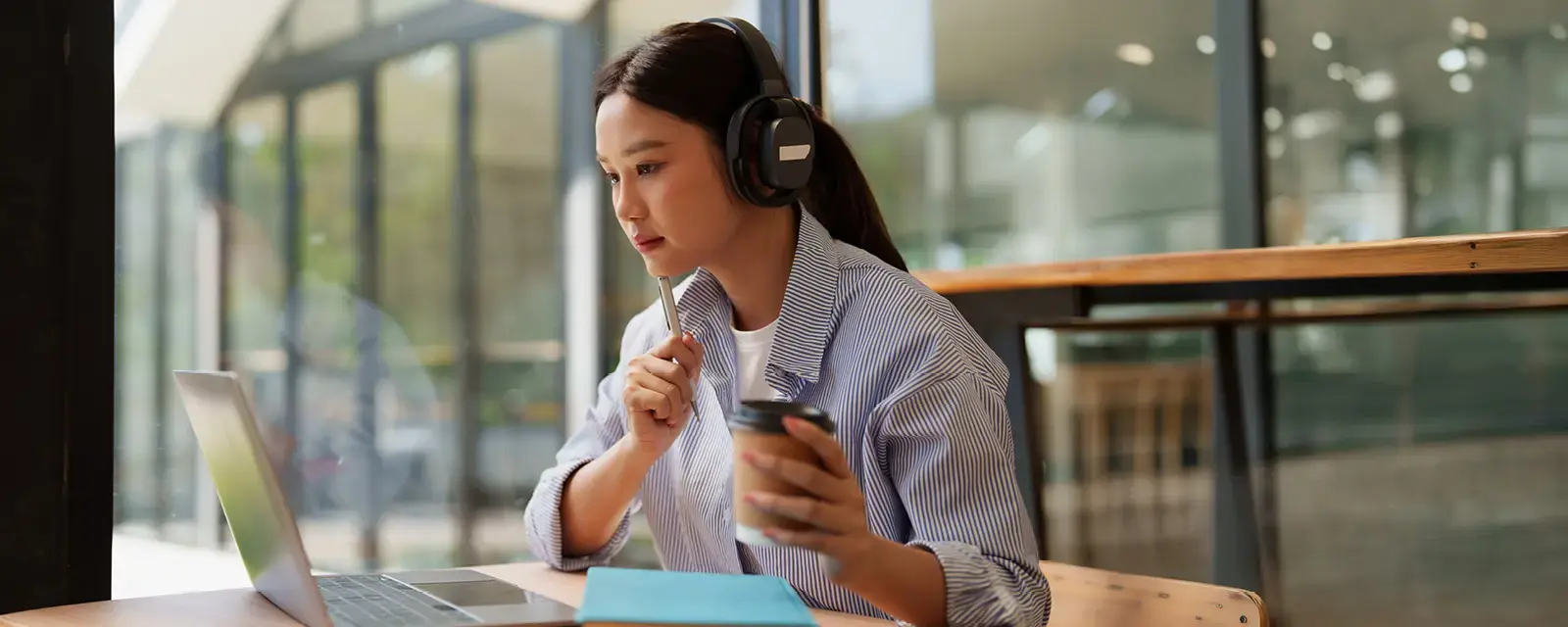 Young woman wearing headphones, holding a pen and to-go coffee, looking a laptop.