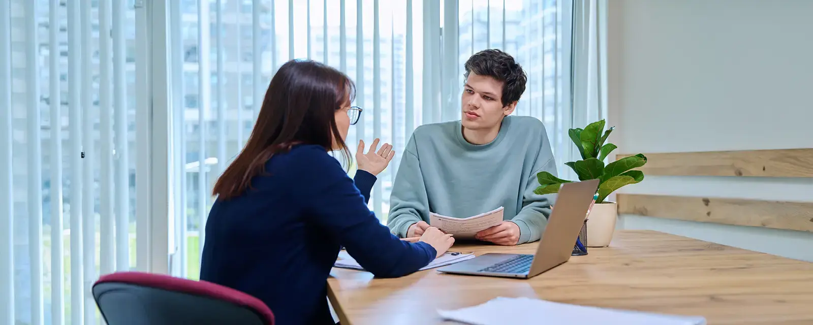 "Middle aged woman, a teacher and mentor, advising male student sitting in a  office building by the window.   "