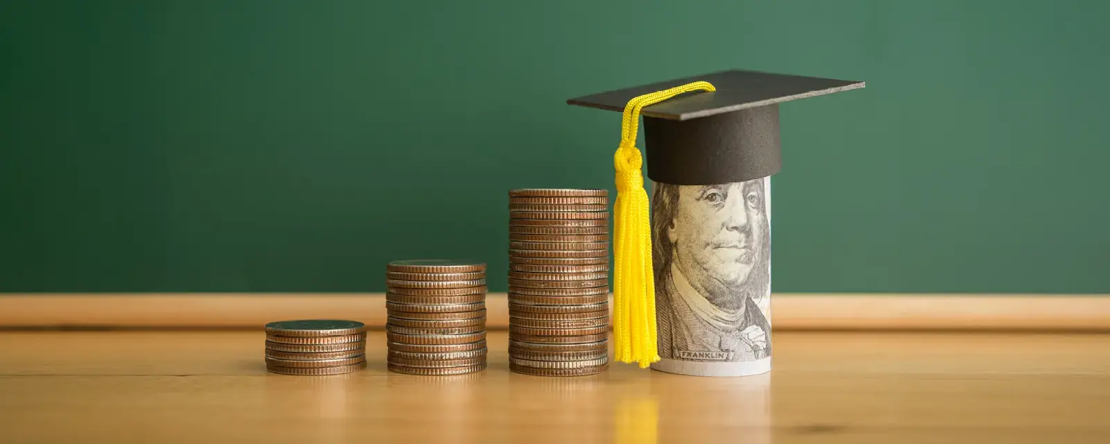 Two stacks of coins next to a rolled up dollar bill showing Ben Franklin's face with a small mortarboard on top.
