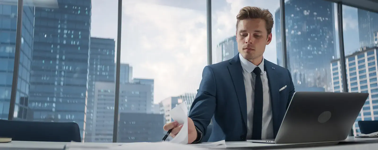 Man wearing a suit, working at a desk with windows and a cityscape behind him.