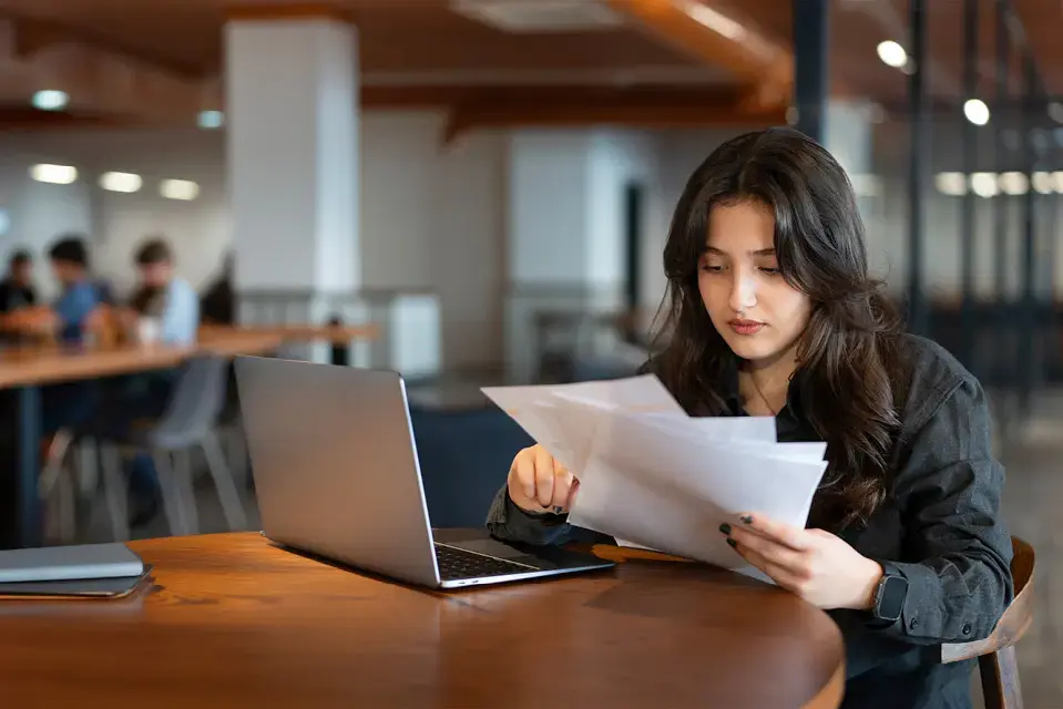 Young woman works with laptop in cafe and examines financial report papers.