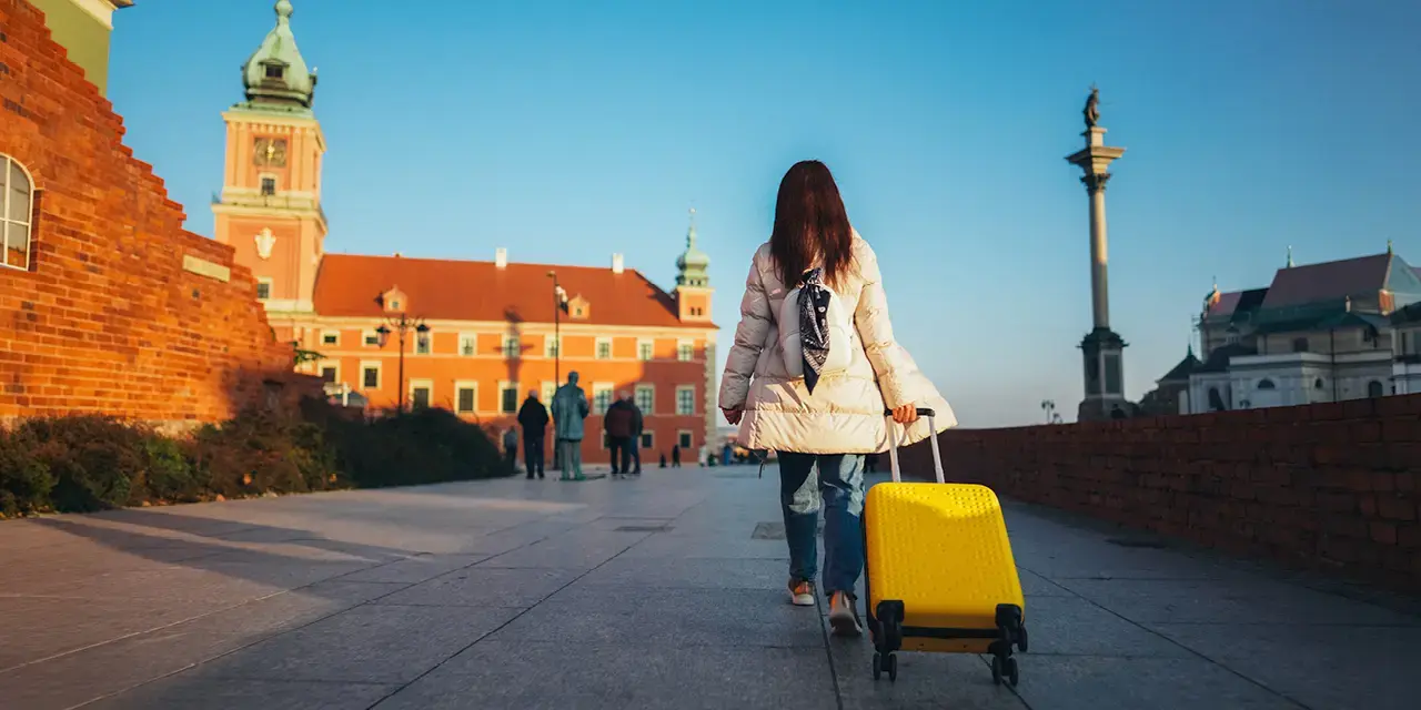 A woman with a white coat pulls a yellow suitcase on wheels next to a brick wall. 