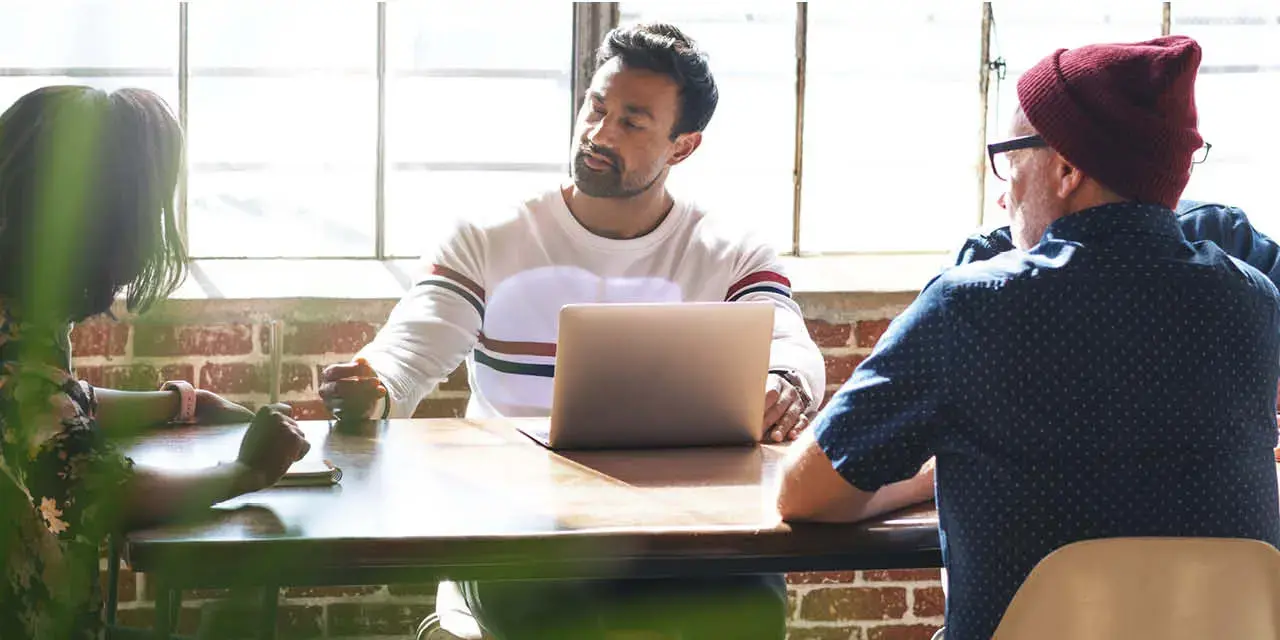 Diverse group of people having a meeting at table with a laptop. They are engaged in discussion, sitting in large windows room. Diverse people discussion at table in modern room. Meeting and discuss