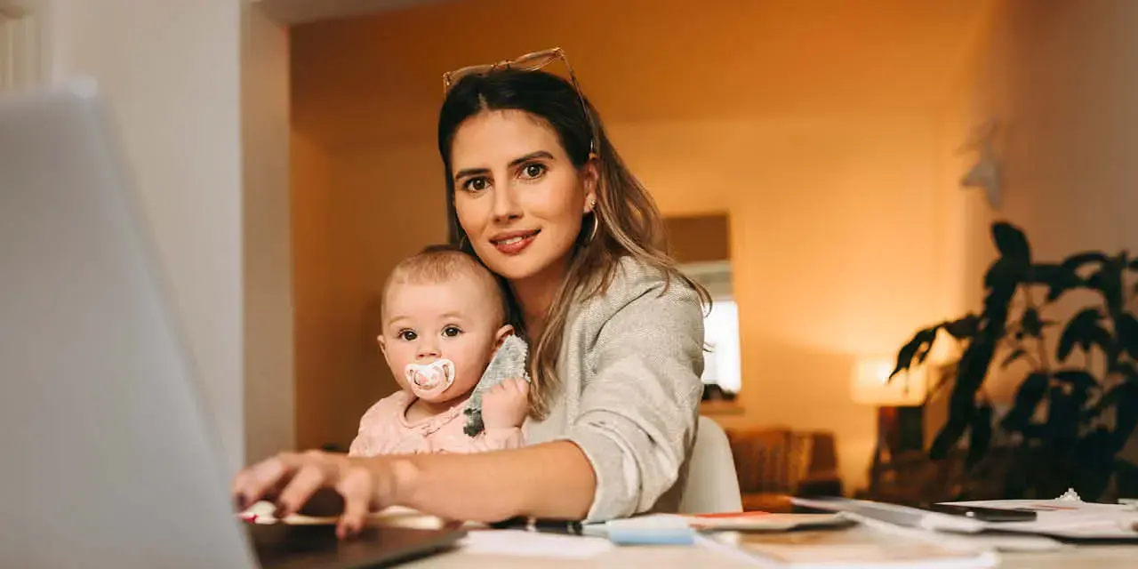 Single mom typing on a laptop while working in her home office. Multitasking mom working on a new creative project at her desk. Female interior designer carrying her baby on her laps.