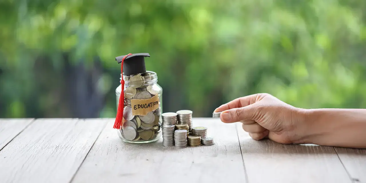 Stacks of coins next to a glass jar filled with coins labled 'Education'. There is a graduation cap on top of the jar.