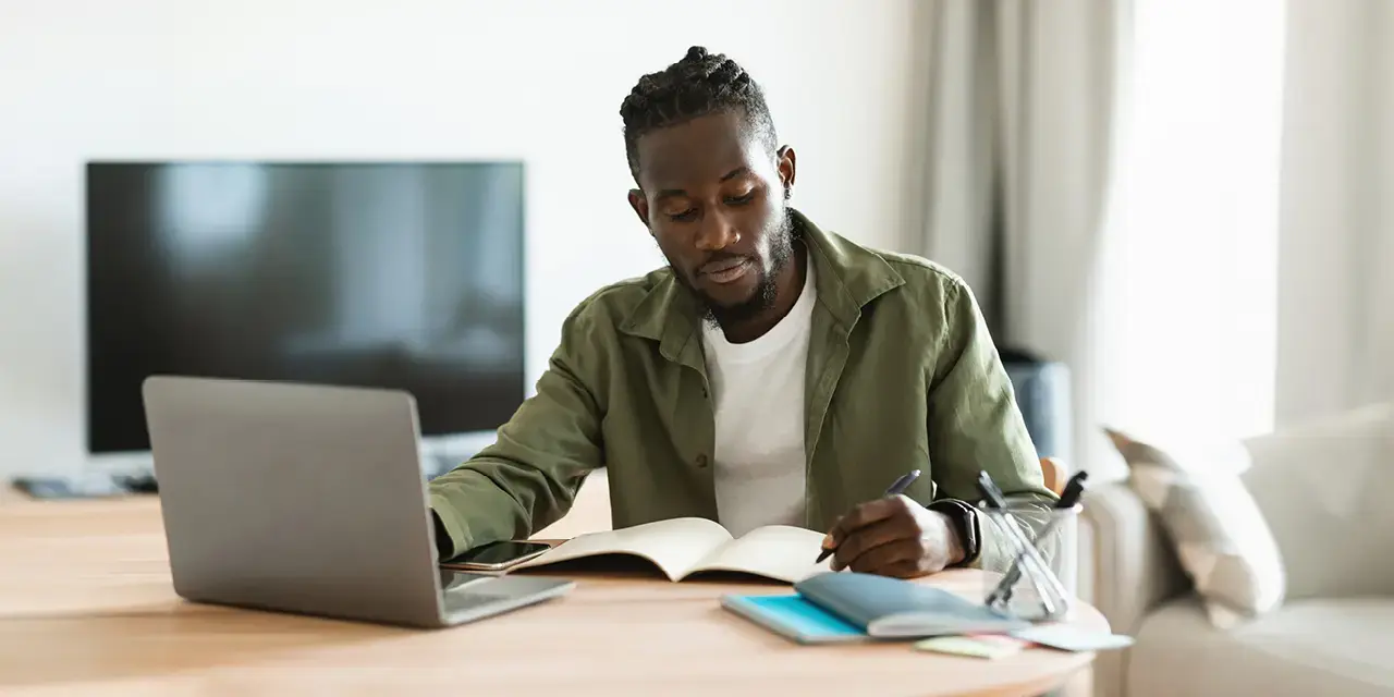 "Focused African-American man sitting at desk working on laptop and taking notes in notebook at home.   "