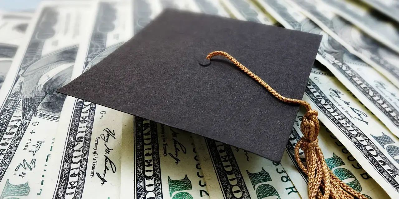 Close up of a black graduation cap, or mortaboard, with gold tassel on top of hundred dollar bills