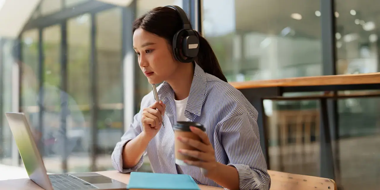 Young woman wearing headphones, holding a pen and to-go coffee, looking a laptop.