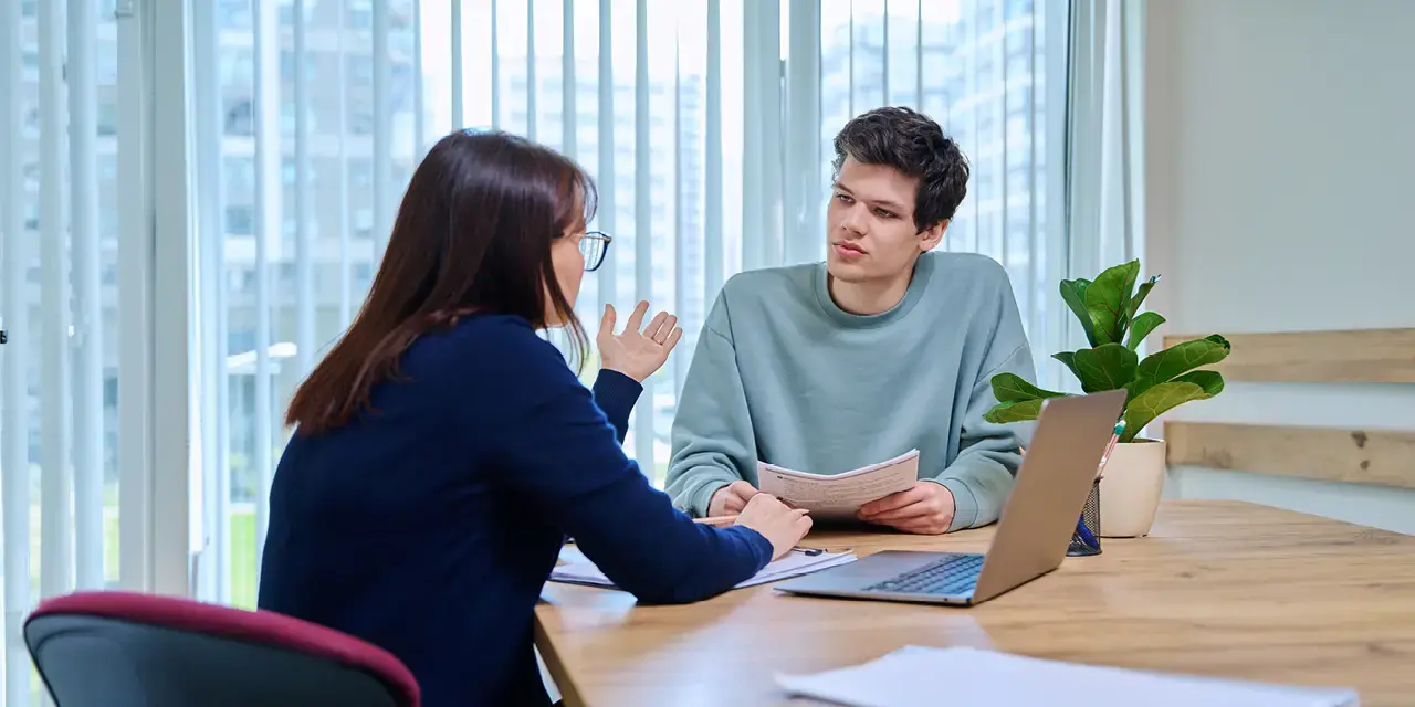"Middle aged woman, a teacher and mentor, advising male student sitting in a  office building by the window.   "