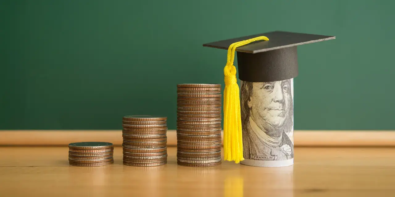 Two stacks of coins next to a rolled up dollar bill showing Ben Franklin's face with a small mortarboard on top.
