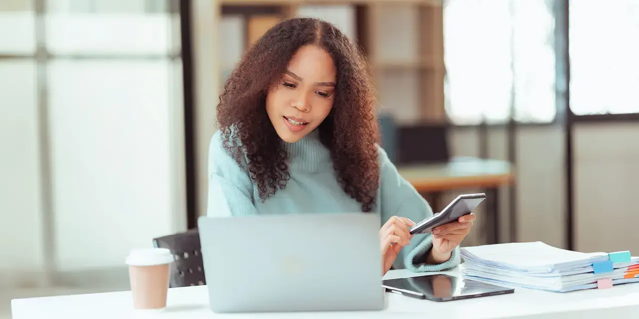 Young woman holding a calculator, looking at a laptop.