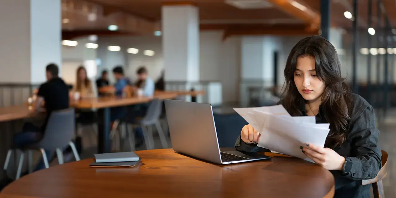 Young woman works with laptop in cafe and examines financial report papers.