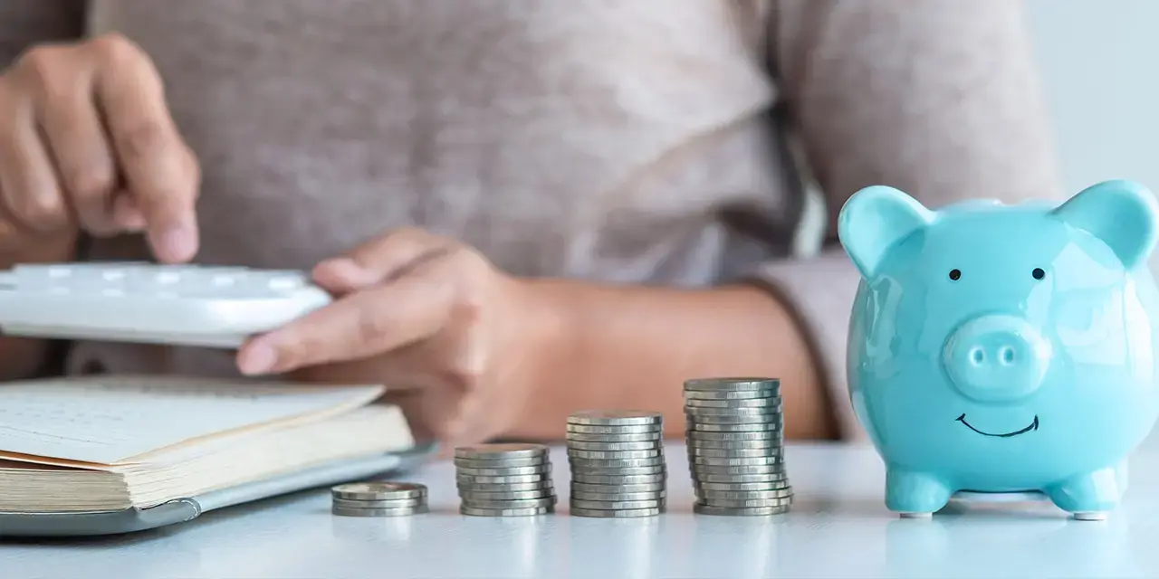 Close-up of a person using a calculator with a piggy bank and coins in the foreground. 