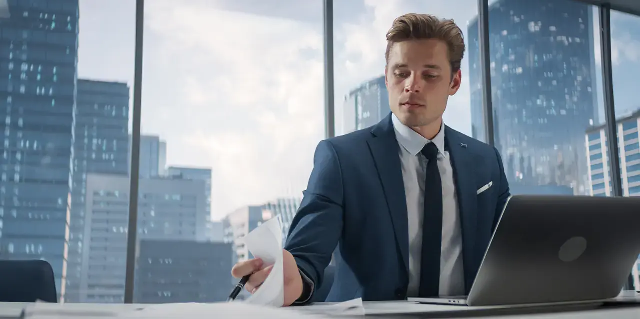 Man wearing a suit, working at a desk with windows and a cityscape behind him.