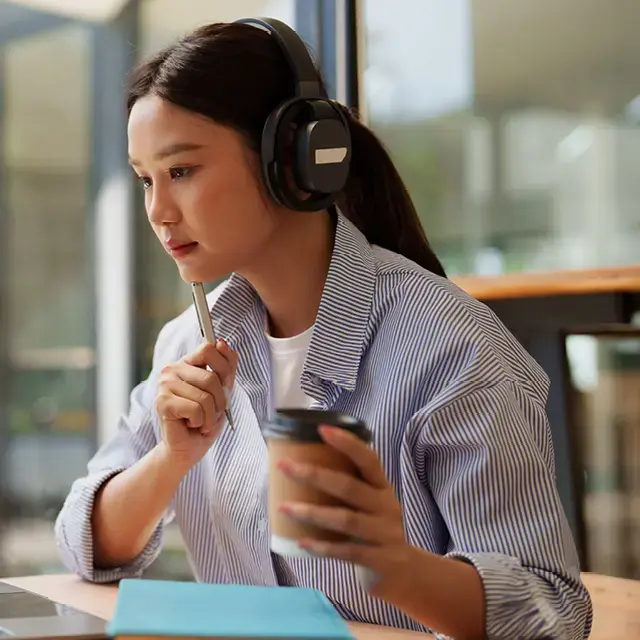 Young woman wearing headphones, holding a pen and to-go coffee, looking a laptop.