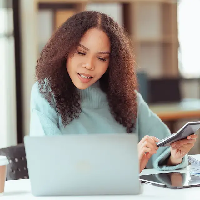 Young woman holding a calculator, looking at a laptop.
