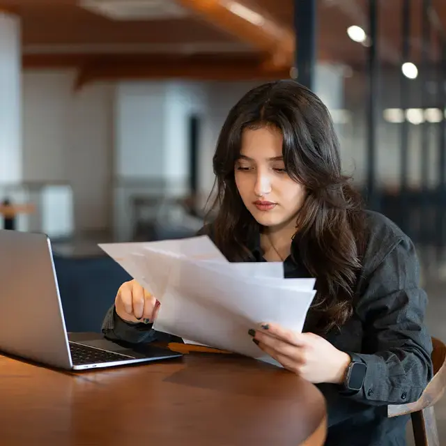Young woman works with laptop in cafe and examines financial report papers.