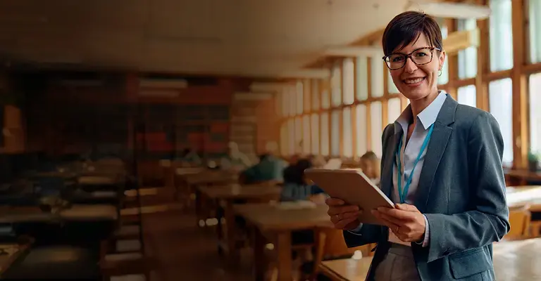 Happy high school teacher using digital tablet in the classroom and looking at camera. Her students are in the background.