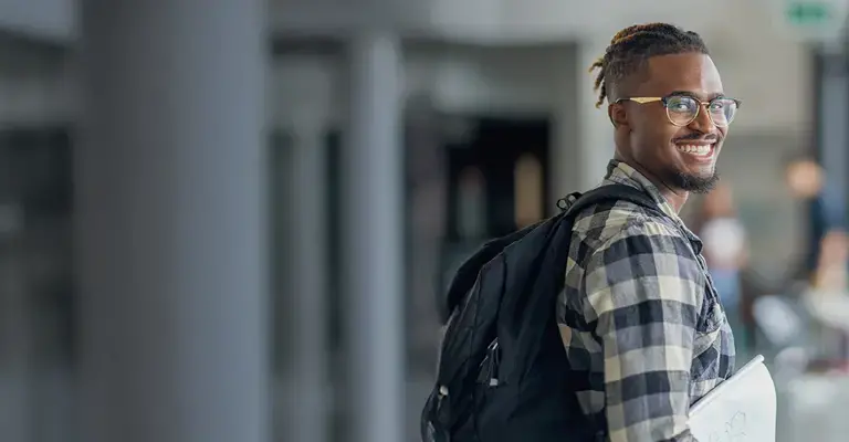 A male University student of African decent, is seen walking to class. He is dressed casually and has his backpack slung over her shoulders as he smiles