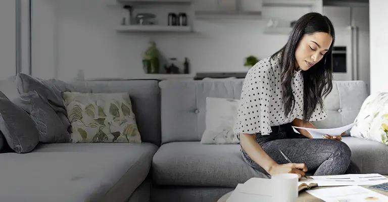 Young woman sitting on a sofa at home analyzing documents and taking notes. There is a digital tablet on a table in front of her. 