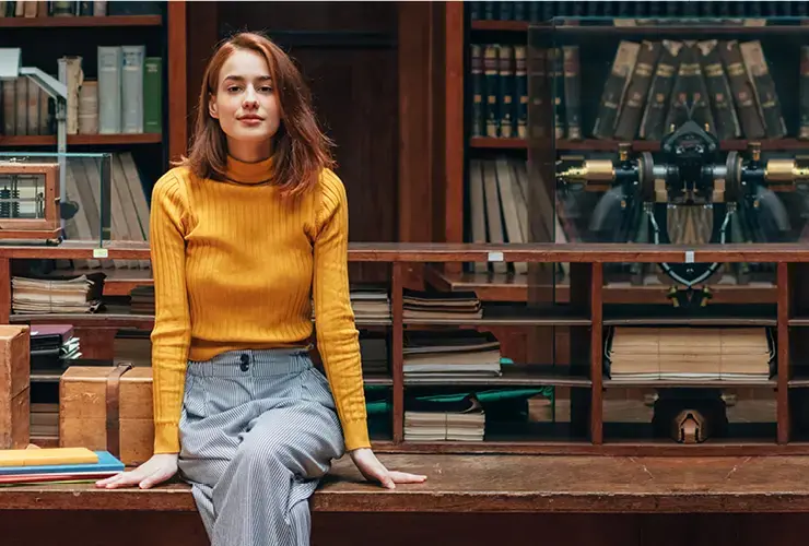 Woman sitting on a library table, with her hands on the table, looking at the camera.