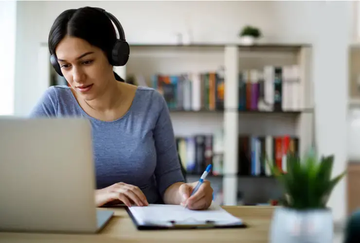 Student wearing headphones, looking at a laptop, and writing notes