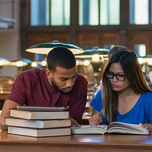 Two students sitting at a library table with open books in front of them.