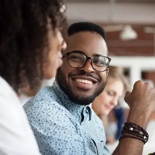 Man wearing glasses and a blue print shirt smiles at the person seated next to him.