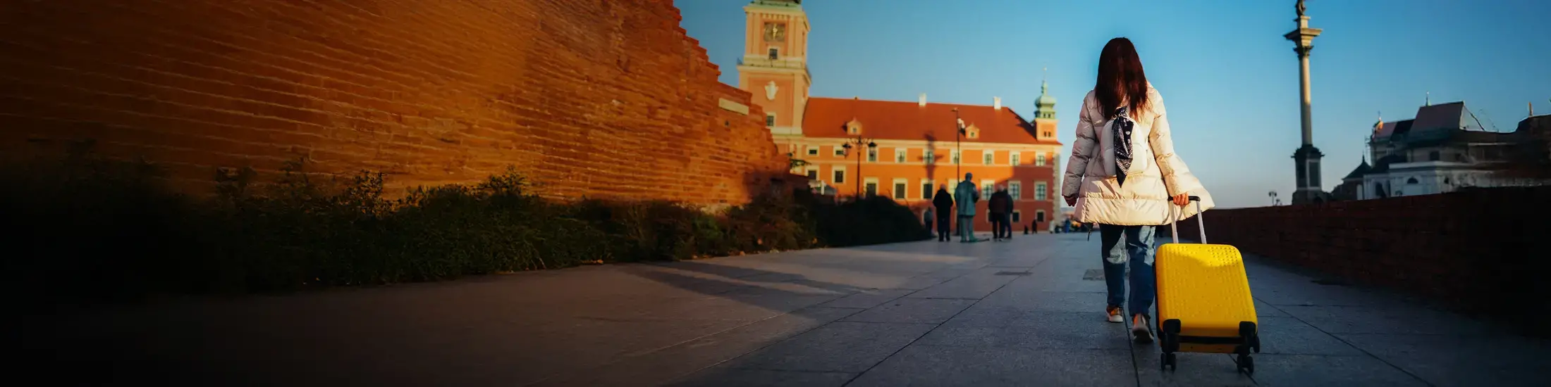 A woman with a white coat pulls a yellow suitcase on wheels next to a brick wall.