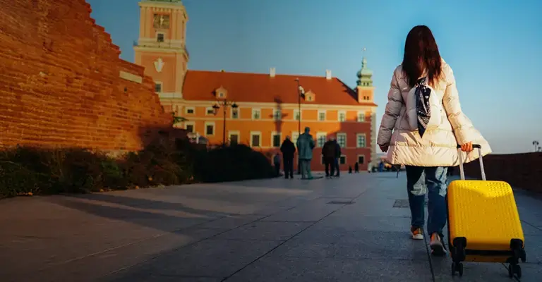 A woman with a white coat pulls a yellow suitcase on wheels next to a brick wall.