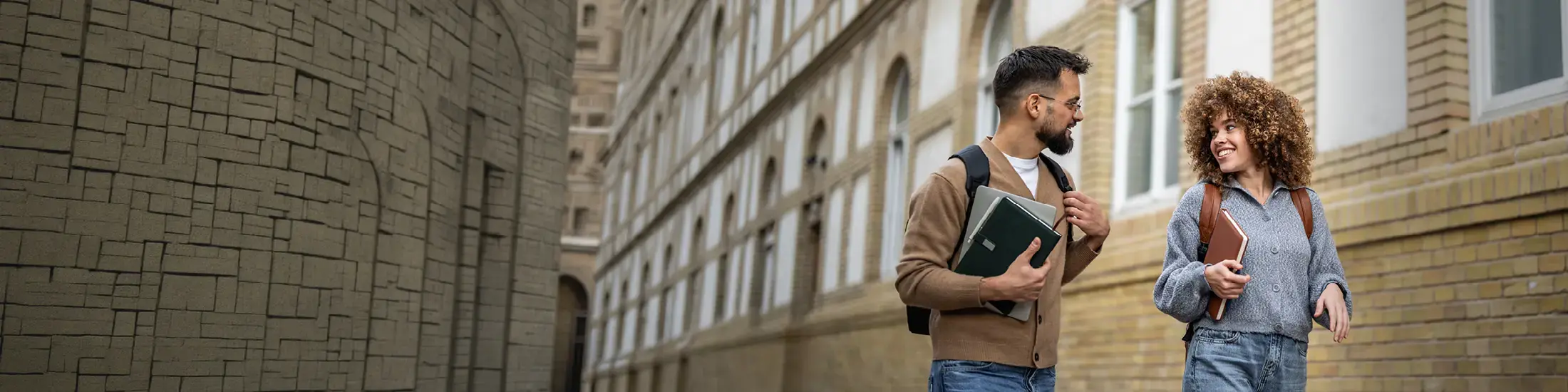 Two young adult students are walking and talking outside of a brick university building, carrying books and backpacks, enjoying their time on campus