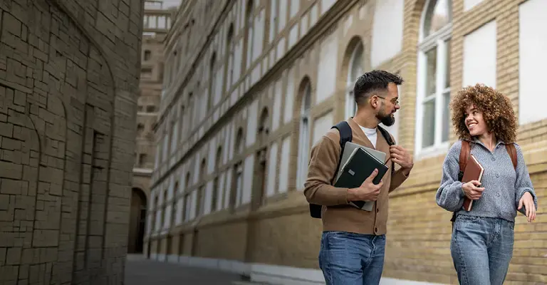 Two young adult students are walking and talking outside of a brick university building, carrying books and backpacks, enjoying their time on campus