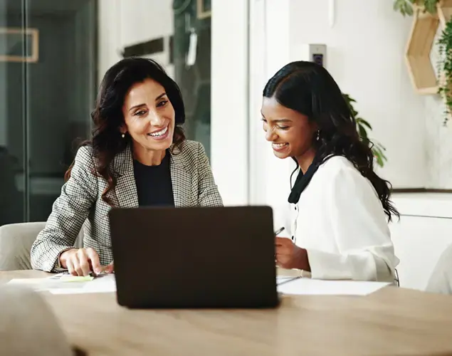 Two professional woman sitting side by side at a table looking at a laptop