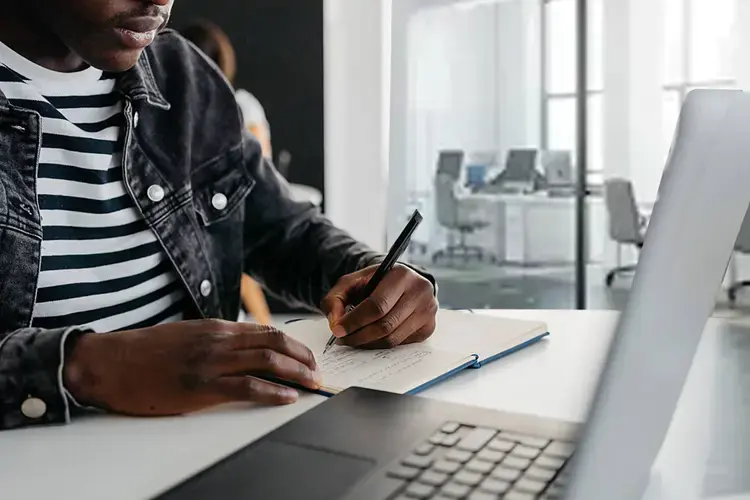 Person writing in a notebook next to a laptop in a bright office, with desks and computers in the background.