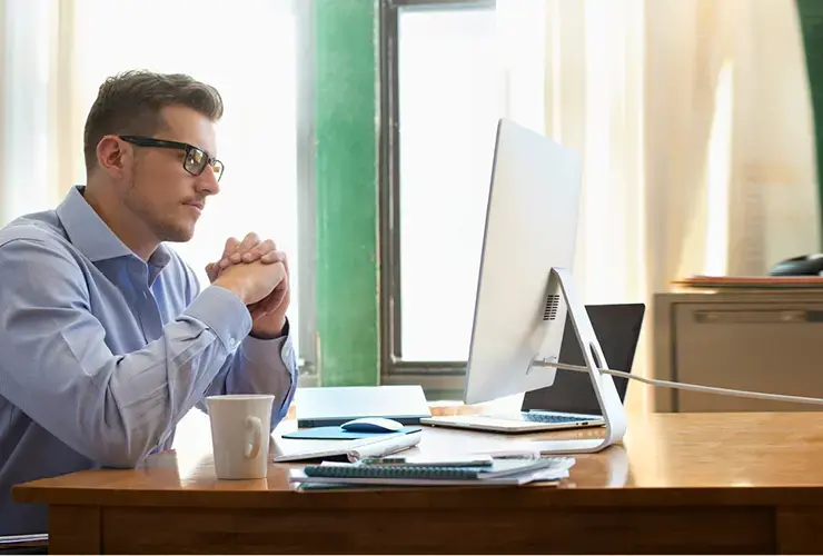 Person wearing glasses and a blue shirt at an iMac, hands clasped, with a coffee mug, printer, brick wall, and desk items.