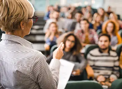 Presenter at the front of a classroom holding papers and gesturing toward an audience seated in lecture hall rows.